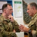 Two military officers in MultiCam/OCP camouflage uniforms are engaged in a focused conversation. The officer on the left, identified by a name tape reading 'ANDERSON' and wearing four stars indicating a General rank, is listening attentively with hand raised to chin. The officer on the right wears a Polish flag patch and a unit patch, suggesting a multinational military meeting or briefing.