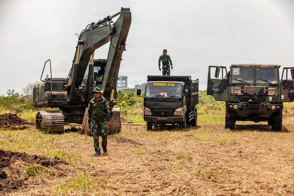 Military personnel in camouflage uniforms are conducting an engineering or construction operation in an open field. The scene includes heavy earth-moving equipment and military trucks, suggesting a land-clearing or infrastructure development exercise. The text 'YONZIKON 12/KJ' visible on one truck indicates Indonesian Army (TNI-AD) Combat Engineer Battalion involvement.