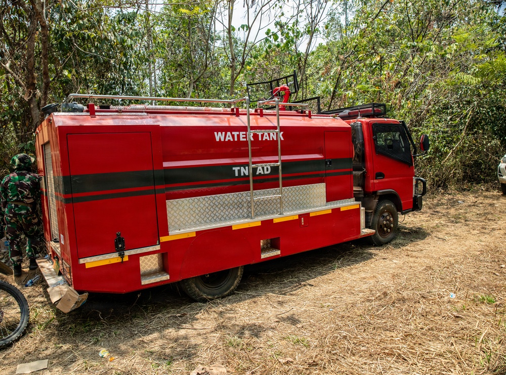 A red water tank truck marked 'TNI AD' (Indonesian Army) is parked in a dry, wooded area. The vehicle appears to be deployed for firefighting or water supply operations in a forested environment. A person in military camouflage uniform is visible on the left side of the vehicle.
