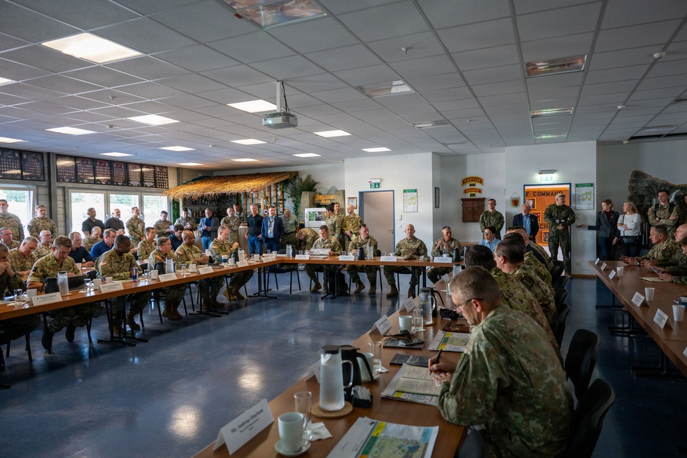 A large military briefing or conference is taking place in a spacious meeting room, with numerous personnel in camouflage uniforms seated around rectangular tables arranged in a U-shape. Additional attendees, including some civilians, are standing along the walls. The room features military insignia, plaques, and unit patches on the walls, suggesting a formal command-level gathering.