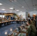 A large military briefing or conference is taking place in a spacious meeting room, with numerous personnel in camouflage uniforms seated around rectangular tables arranged in a U-shape. Additional attendees, including some civilians, are standing along the walls. The room features military insignia, plaques, and unit patches on the walls, suggesting a formal command-level gathering.
