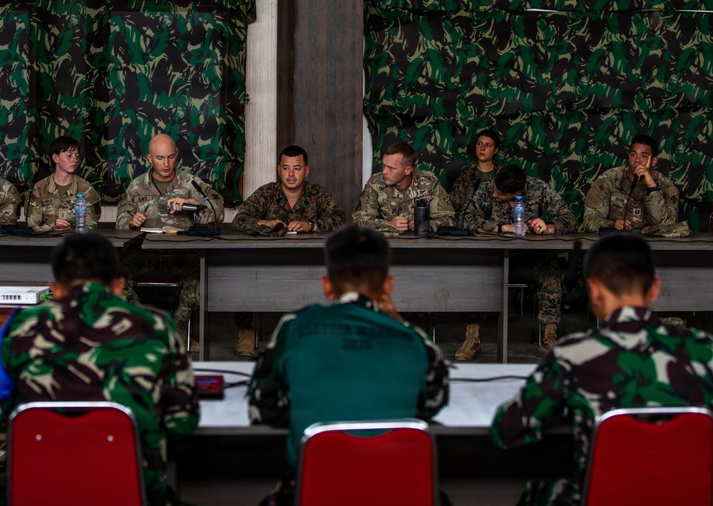 A military briefing or meeting is taking place between two groups of soldiers in a room decorated with camouflage-patterned fabric on the walls. One group, wearing MultiCam/OCP uniforms consistent with US military dress, is seated at a long table facing the camera, while another group in a different camouflage pattern (appearing to be Indonesian TNI-style woodland camouflage) is seated with their backs to the camera. The setting suggests a joint military exercise or bilateral training event.