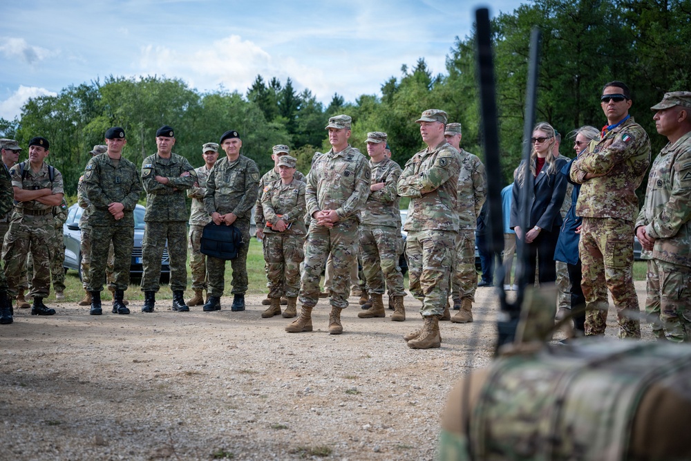 A multinational military gathering is taking place outdoors at what appears to be a training area or military installation. Personnel in various camouflage uniforms, including US Army OCP pattern and other NATO-style uniforms, are assembled in a semicircle, apparently receiving a briefing or demonstration. Two civilian individuals are also present among the military personnel.