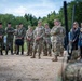 A multinational military gathering is taking place outdoors at what appears to be a training area or military installation. Personnel in various camouflage uniforms, including US Army OCP pattern and other NATO-style uniforms, are assembled in a semicircle, apparently receiving a briefing or demonstration. Two civilian individuals are also present among the military personnel.