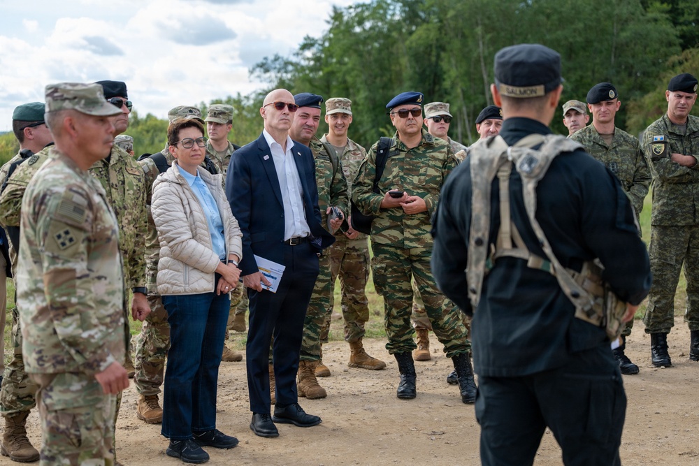 A group of military personnel from multiple nations and at least two civilians are gathered outdoors on a dirt path, appearing to attend a briefing or demonstration. The scene suggests a multinational military exercise or official visit, with personnel wearing various camouflage patterns and uniforms. A figure in black tactical gear with a name tag reading 'SALMON' is facing the group, likely conducting the briefing.