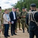 A group of military personnel from multiple nations and at least two civilians are gathered outdoors on a dirt path, appearing to attend a briefing or demonstration. The scene suggests a multinational military exercise or official visit, with personnel wearing various camouflage patterns and uniforms. A figure in black tactical gear with a name tag reading 'SALMON' is facing the group, likely conducting the briefing.