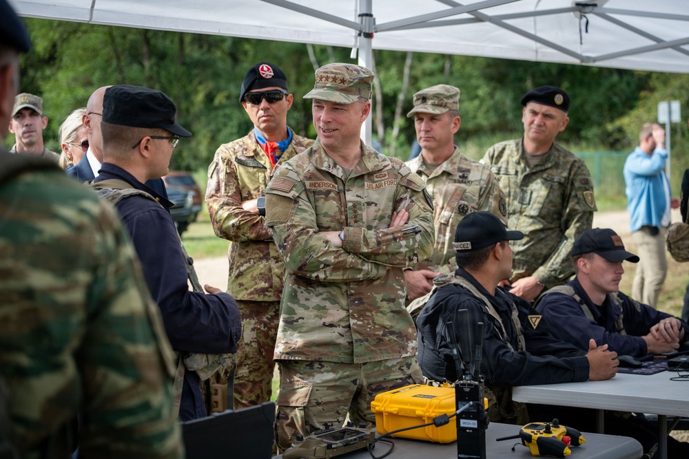 A group of military personnel from multiple nations are gathered under a white canopy tent at what appears to be an outdoor command post or exercise briefing area. A US Air Force officer with three stars (Lieutenant General) and the name tape 'ANDERSON' is prominently featured in the center, standing with arms crossed while engaged in conversation. Personnel are seated at tables with communications equipment including radios and yellow Pelican cases.