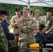 A group of military personnel from multiple nations are gathered under a white canopy tent at what appears to be an outdoor command post or exercise briefing area. A US Air Force officer with three stars (Lieutenant General) and the name tape 'ANDERSON' is prominently featured in the center, standing with arms crossed while engaged in conversation. Personnel are seated at tables with communications equipment including radios and yellow Pelican cases.