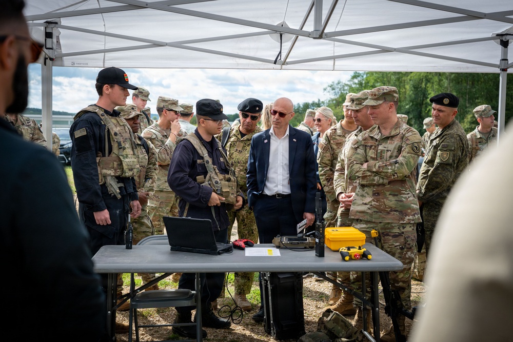 A group of military personnel in camouflage uniforms and a civilian in a suit gather under a white canopy tent for what appears to be a equipment demonstration or briefing. A table in the foreground holds various items including a laptop, a yellow hard case, and other technical equipment. The setting appears to be an outdoor military exercise or demonstration area.