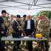 A group of military personnel in camouflage uniforms and a civilian in a suit gather under a white canopy tent for what appears to be a equipment demonstration or briefing. A table in the foreground holds various items including a laptop, a yellow hard case, and other technical equipment. The setting appears to be an outdoor military exercise or demonstration area.