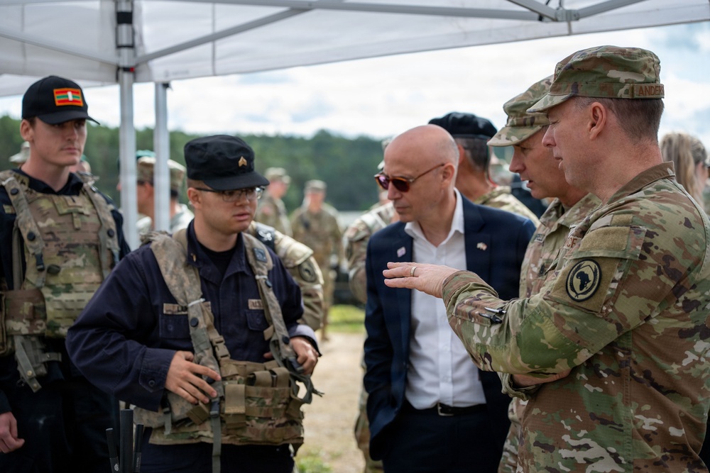 A military briefing or discussion is taking place outdoors under a white canopy tent. A senior military officer in OCP (Operational Camouflage Pattern) uniform, with a name tape reading 'ANDER' and a USAFRICOM patch, appears to be briefing a civilian in a suit wearing an American flag lapel pin alongside other military personnel. Personnel in mixed uniforms and tactical gear, including black caps and plate carriers, are present.