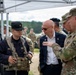 A military briefing or discussion is taking place outdoors under a white canopy tent. A senior military officer in OCP (Operational Camouflage Pattern) uniform, with a name tape reading 'ANDER' and a USAFRICOM patch, appears to be briefing a civilian in a suit wearing an American flag lapel pin alongside other military personnel. Personnel in mixed uniforms and tactical gear, including black caps and plate carriers, are present.