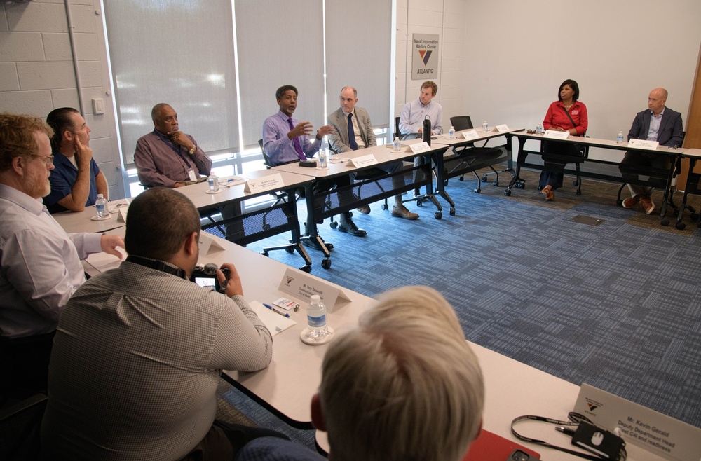 A formal meeting or briefing is taking place at what appears to be the Naval Information Warfare Center Atlantic, as indicated by the banner visible in the background. Participants are seated around conference tables arranged in a U-shape, with name placards identifying attendees. One individual in a purple shirt appears to be actively speaking or presenting to the group.