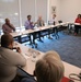 A formal meeting or briefing is taking place at what appears to be the Naval Information Warfare Center Atlantic, as indicated by the banner visible in the background. Participants are seated around conference tables arranged in a U-shape, with name placards identifying attendees. One individual in a purple shirt appears to be actively speaking or presenting to the group.
