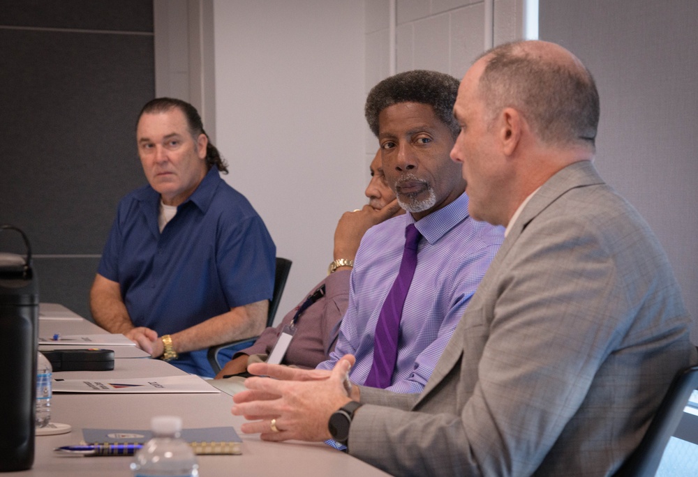 Four men are seated at a conference table in what appears to be a formal meeting or discussion setting. Three men are clearly visible in the foreground, while a fourth is partially visible in the background. The men appear engaged in conversation, with at least one actively speaking.