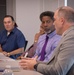 Four men are seated at a conference table in what appears to be a formal meeting or discussion setting. Three men are clearly visible in the foreground, while a fourth is partially visible in the background. The men appear engaged in conversation, with at least one actively speaking.