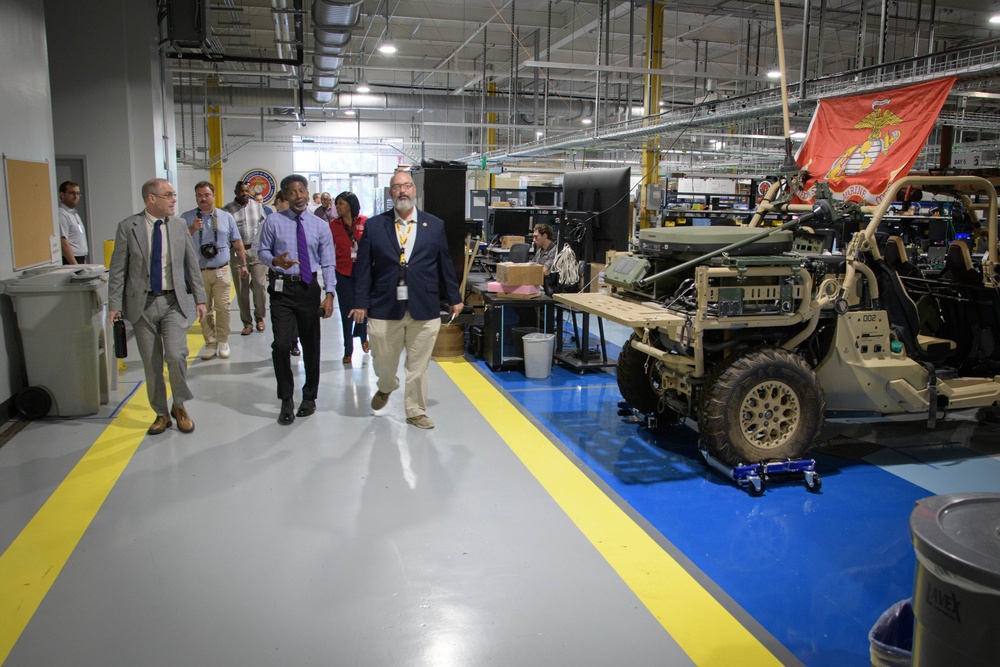 A group of civilians and officials are touring what appears to be a U.S. Marine Corps military maintenance or integration facility. The facility contains military tactical vehicles under assembly or maintenance, with a U.S. Marine Corps flag prominently displayed. Visitors wearing business attire and identification badges are being guided through the workspace.