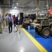 A group of civilians and officials are touring what appears to be a U.S. Marine Corps military maintenance or integration facility. The facility contains military tactical vehicles under assembly or maintenance, with a U.S. Marine Corps flag prominently displayed. Visitors wearing business attire and identification badges are being guided through the workspace.