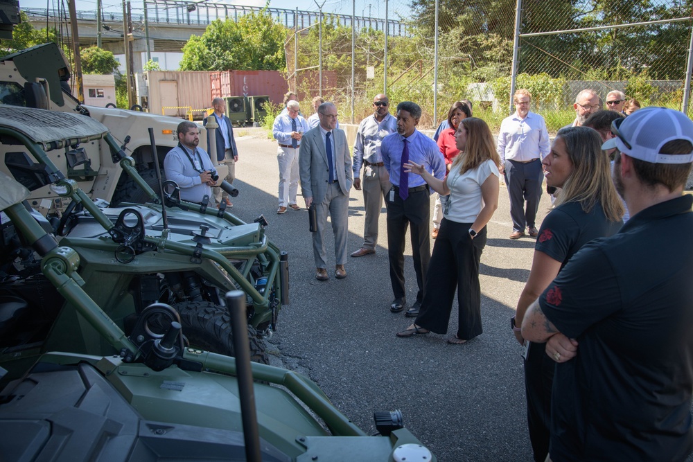 A group of approximately 15-20 civilians and officials are gathered outdoors around military ground vehicles, appearing to be on an organized tour or briefing. A woman in a white top appears to be presenting or explaining something to the group near what looks like a light tactical military vehicle painted in olive green. The setting appears to be a military installation or depot with fencing, shipping containers, and an overpass visible in the background.
