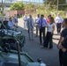 A group of approximately 15-20 civilians and officials are gathered outdoors around military ground vehicles, appearing to be on an organized tour or briefing. A woman in a white top appears to be presenting or explaining something to the group near what looks like a light tactical military vehicle painted in olive green. The setting appears to be a military installation or depot with fencing, shipping containers, and an overpass visible in the background.