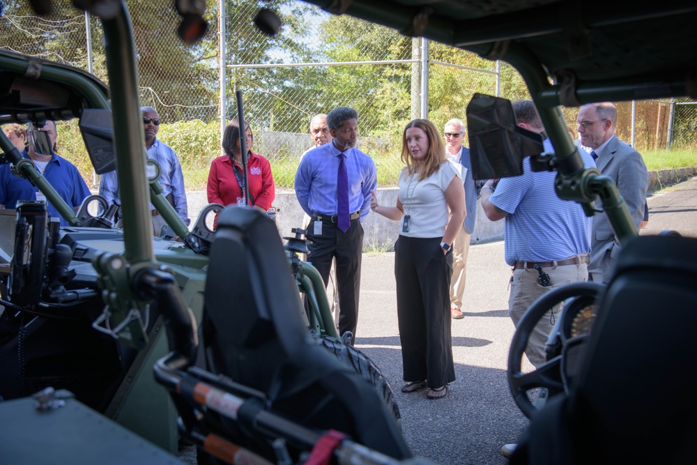 A group of approximately 8-10 civilians in business casual attire are gathered outdoors around a military-style green utility vehicle, likely during a demonstration or briefing. The image is shot from inside or very close to the vehicle, giving a low-angle perspective through the vehicle's frame. A woman in a white top and black pants appears to be presenting or explaining something to the group.