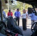 A group of approximately 8-10 civilians in business casual attire are gathered outdoors around a military-style green utility vehicle, likely during a demonstration or briefing. The image is shot from inside or very close to the vehicle, giving a low-angle perspective through the vehicle's frame. A woman in a white top and black pants appears to be presenting or explaining something to the group.