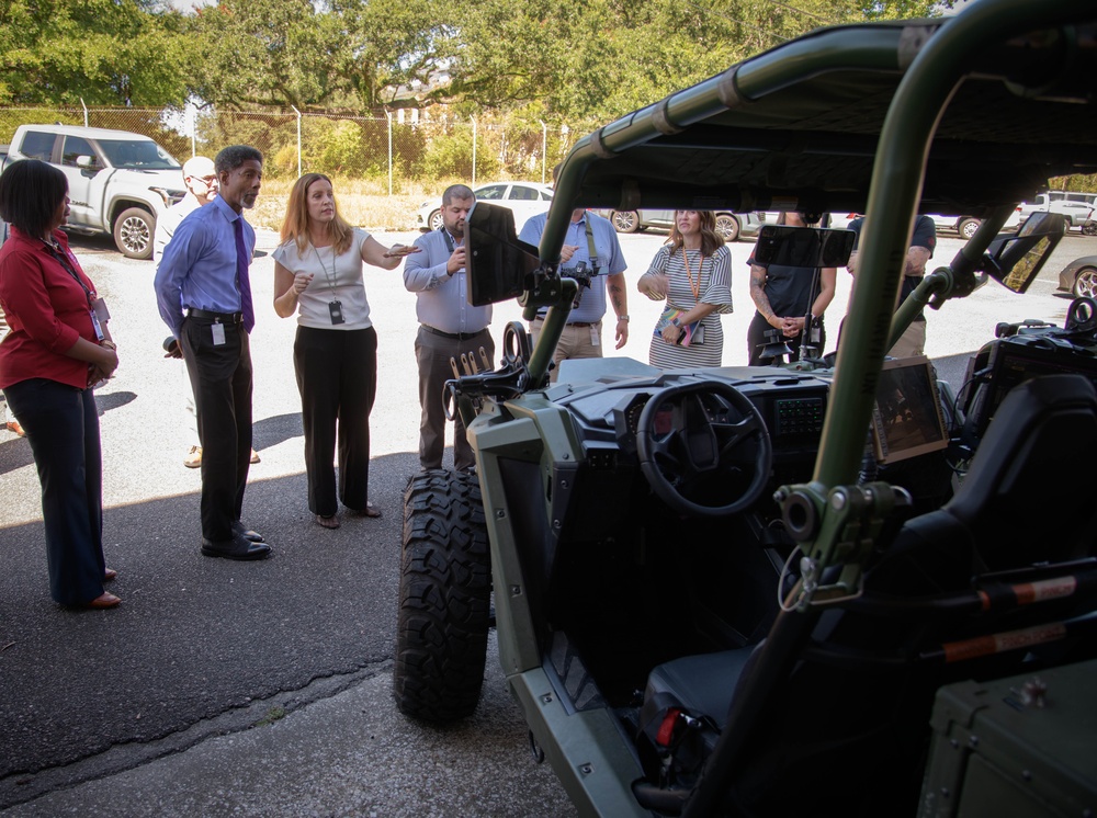 A group of approximately 8-10 people, appearing to be professionals or officials, are gathered in a parking lot around a military-style utility vehicle (likely an LSUTV or similar tactical UTV) painted in olive drab green. A woman in business casual attire appears to be presenting or briefing the group about the vehicle. The setting suggests a demonstration or media event at a government or military facility.