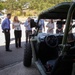 A group of approximately 8-10 people, appearing to be professionals or officials, are gathered in a parking lot around a military-style utility vehicle (likely an LSUTV or similar tactical UTV) painted in olive drab green. A woman in business casual attire appears to be presenting or briefing the group about the vehicle. The setting suggests a demonstration or media event at a government or military facility.