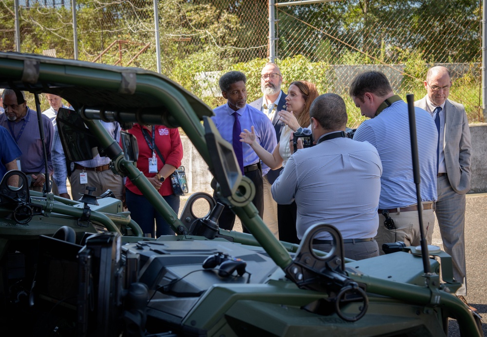 A group of civilians and officials are gathered around military ground vehicles, appearing to be on a formal tour or demonstration. A woman is actively speaking and gesturing, likely briefing or explaining the equipment to the group. At least one person is holding a camera, suggesting media coverage or documentation of the event.