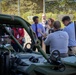 A group of civilians and officials are gathered around military ground vehicles, appearing to be on a formal tour or demonstration. A woman is actively speaking and gesturing, likely briefing or explaining the equipment to the group. At least one person is holding a camera, suggesting media coverage or documentation of the event.