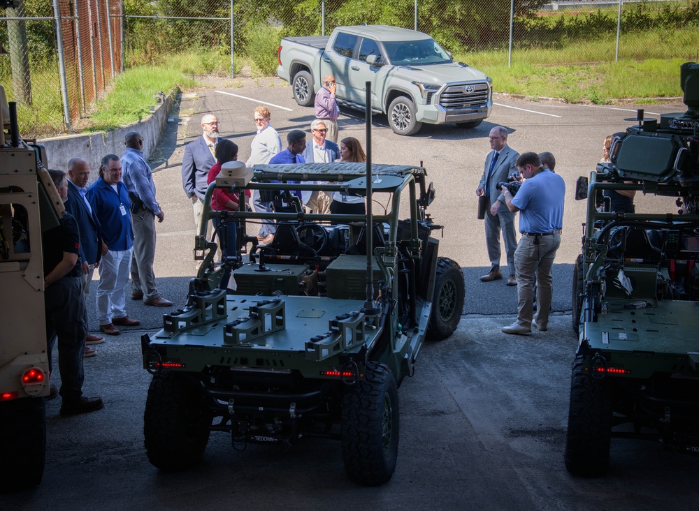 A group of approximately 15-20 civilians and officials in business attire are gathered in a parking lot examining military ground vehicles, likely during a demonstration or briefing event. The vehicles appear to be small tactical unmanned or manned ground vehicles (UGVs) in olive drab/military green, equipped with various sensors and mounting systems. A photographer is documenting the event, and a civilian Toyota Tundra pickup truck is visible in the background.