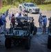 A group of approximately 15-20 civilians and officials in business attire are gathered in a parking lot examining military ground vehicles, likely during a demonstration or briefing event. The vehicles appear to be small tactical unmanned or manned ground vehicles (UGVs) in olive drab/military green, equipped with various sensors and mounting systems. A photographer is documenting the event, and a civilian Toyota Tundra pickup truck is visible in the background.