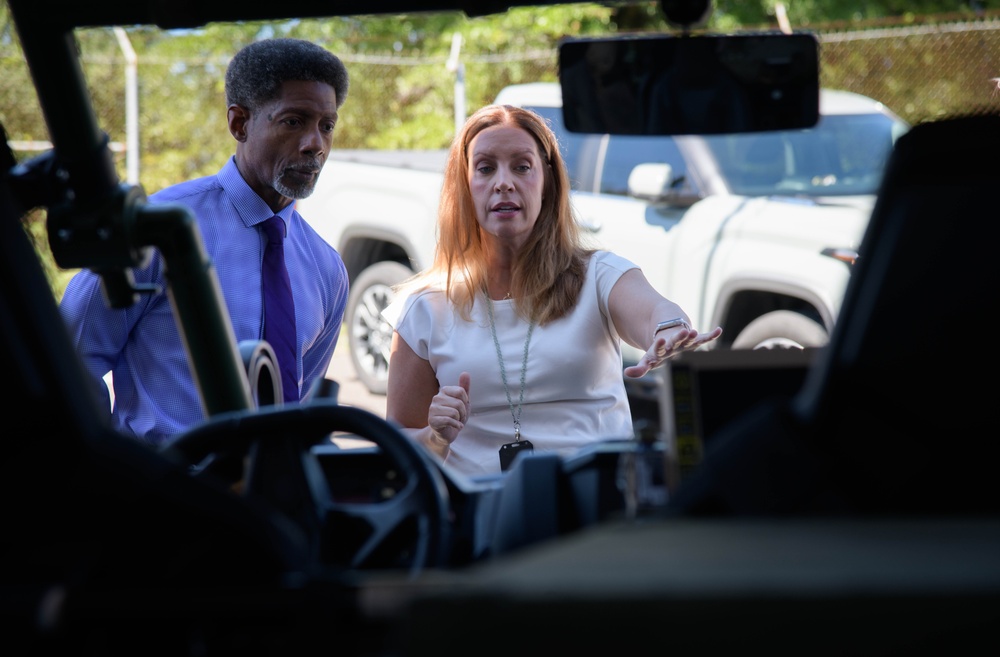 Two individuals, a man and a woman, are engaged in conversation near what appears to be an open utility or off-road vehicle. The woman, wearing a white top and a lanyard with a badge, appears to be explaining or demonstrating something with an outstretched hand. The scene takes place outdoors in a parking or staging area with trucks visible in the background.