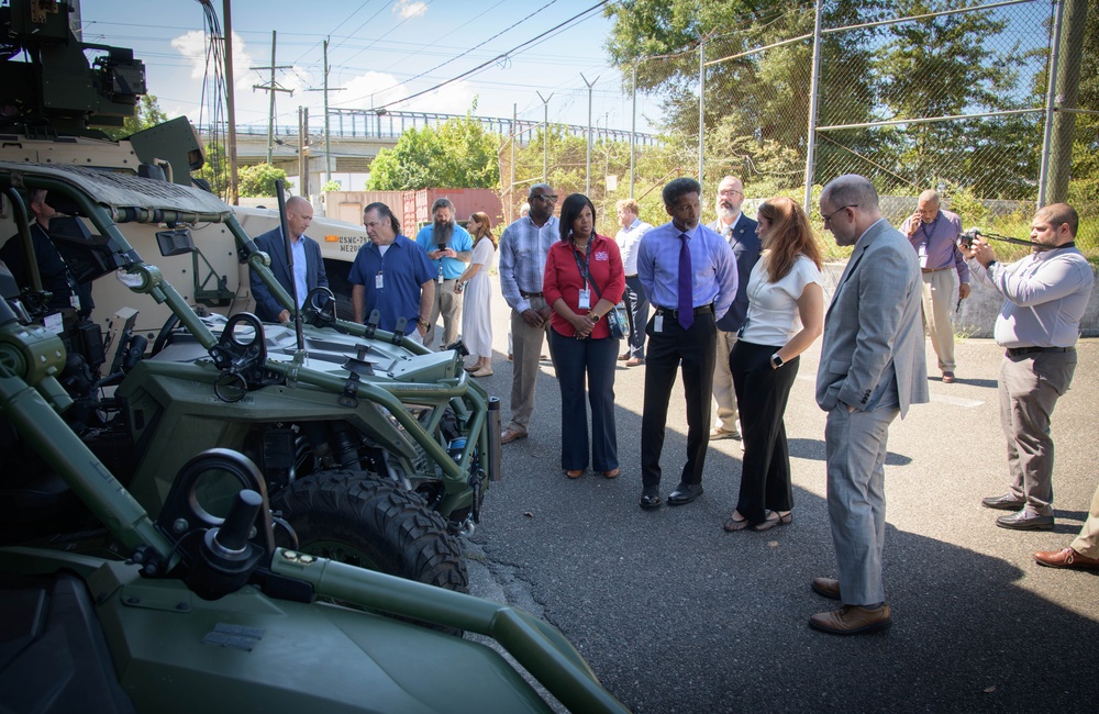 A group of civilians and officials are gathered outdoors examining military ground vehicles, including what appears to be a tactical utility vehicle and a larger armored vehicle. The setting appears to be a demonstration or briefing event at a military or defense facility, indicated by the security fencing and the mix of business-attired attendees with lanyards/badges. Several participants are engaged in conversation while one person photographs the event.