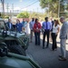 A group of civilians and officials are gathered outdoors examining military ground vehicles, including what appears to be a tactical utility vehicle and a larger armored vehicle. The setting appears to be a demonstration or briefing event at a military or defense facility, indicated by the security fencing and the mix of business-attired attendees with lanyards/badges. Several participants are engaged in conversation while one person photographs the event.
