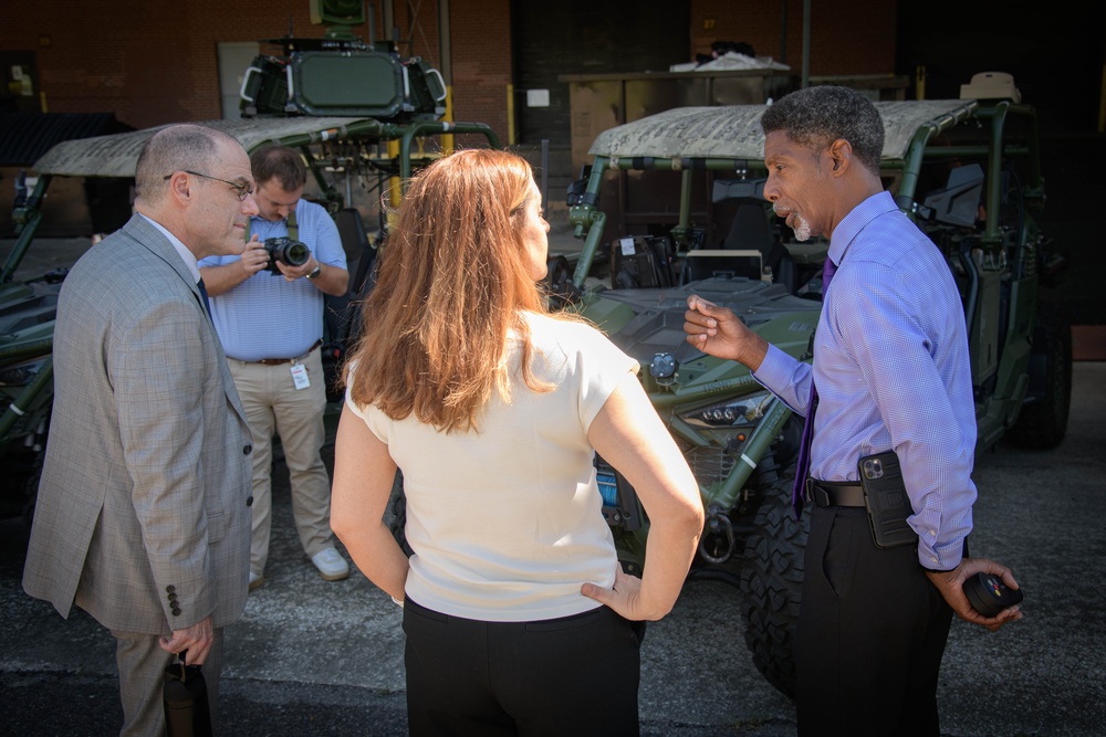 Four individuals are gathered in an outdoor area in front of military-style tactical vehicles, likely at a defense facility or military installation. Three people appear to be engaged in a professional discussion, while a fourth person photographs the scene in the background. The setting and vehicles suggest an official visit, demonstration, or briefing related to military equipment.