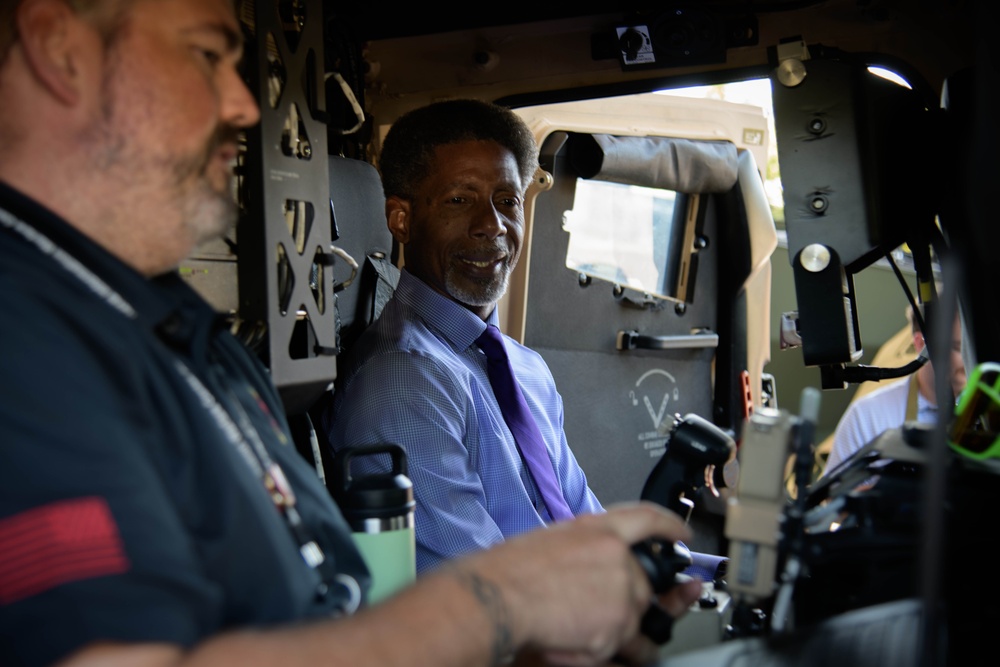Two men are seated inside what appears to be a military vehicle, likely a HMMWV (Humvee) or similar tactical vehicle. The man in the center is wearing a light blue dress shirt with a purple tie and is smiling, while the man on the left is wearing a dark uniform with an American flag patch. They appear to be examining or operating the vehicle's controls and equipment.