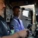 Two men are seated inside what appears to be a military vehicle, likely a HMMWV (Humvee) or similar tactical vehicle. The man in the center is wearing a light blue dress shirt with a purple tie and is smiling, while the man on the left is wearing a dark uniform with an American flag patch. They appear to be examining or operating the vehicle's controls and equipment.