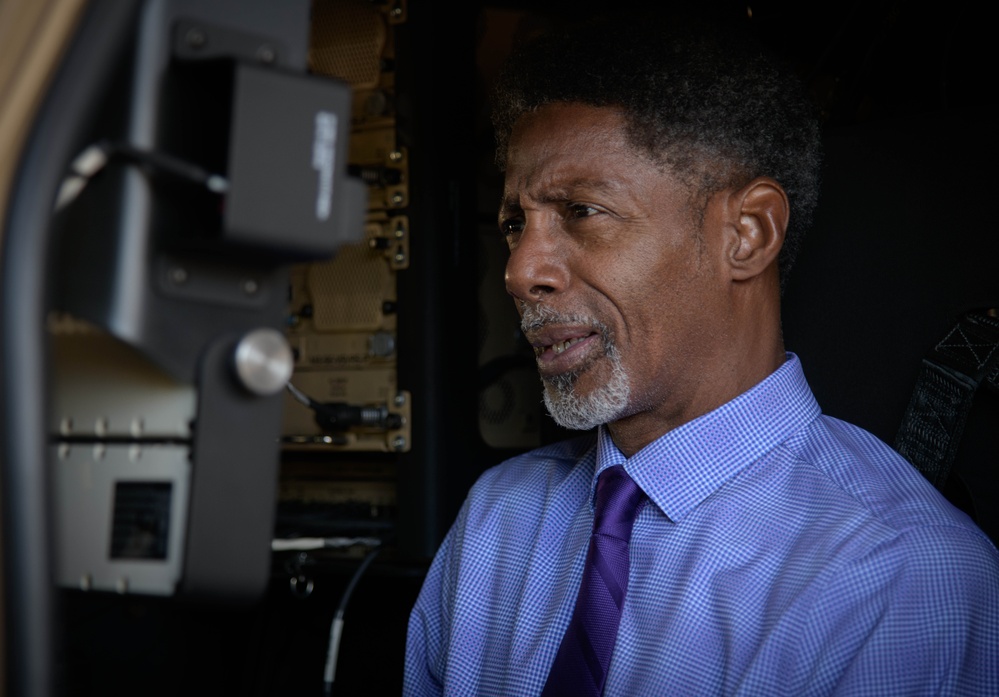 A middle-aged man wearing a blue checkered dress shirt and purple tie is photographed in profile view. He appears to be inside or adjacent to a piece of heavy equipment or military vehicle, with visible mechanical components and wiring panels in the background. The image is a close-up portrait with shallow depth of field.