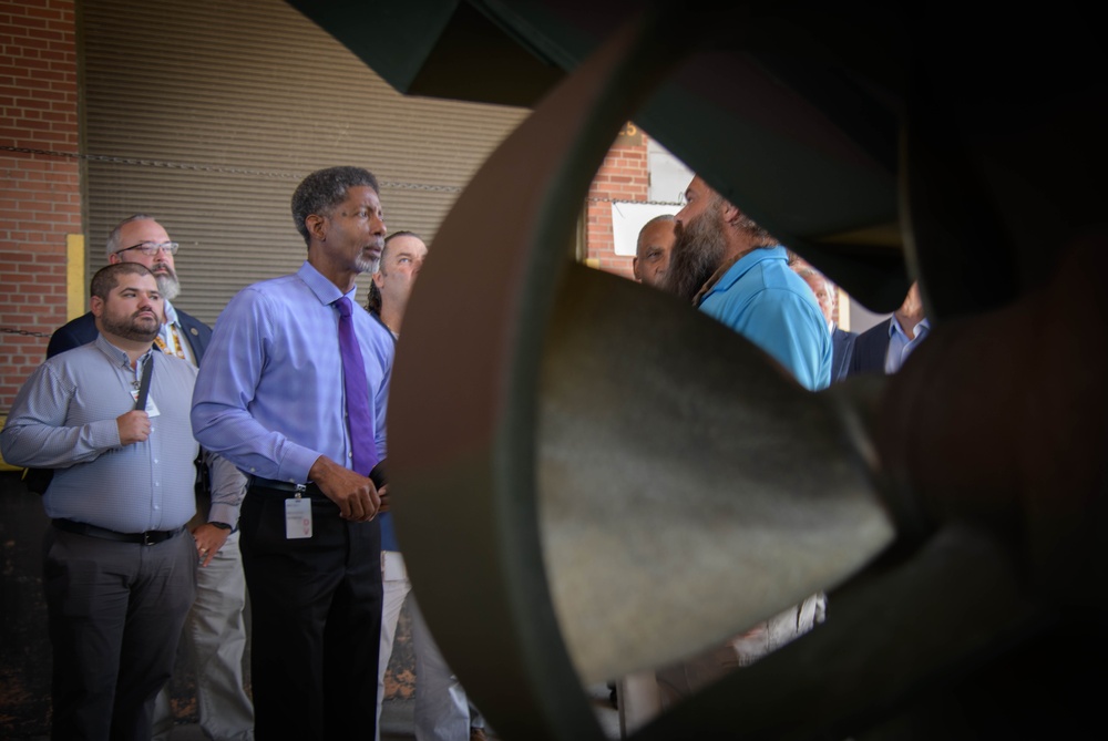 A group of professionals or officials are gathered outdoors near a brick building, appearing to examine or discuss a large piece of industrial or mechanical equipment in the foreground. The central figure, wearing a light blue dress shirt and purple tie with a visible ID badge, appears to be speaking or presenting to the group. The scene suggests a facility tour, inspection, or official visit.