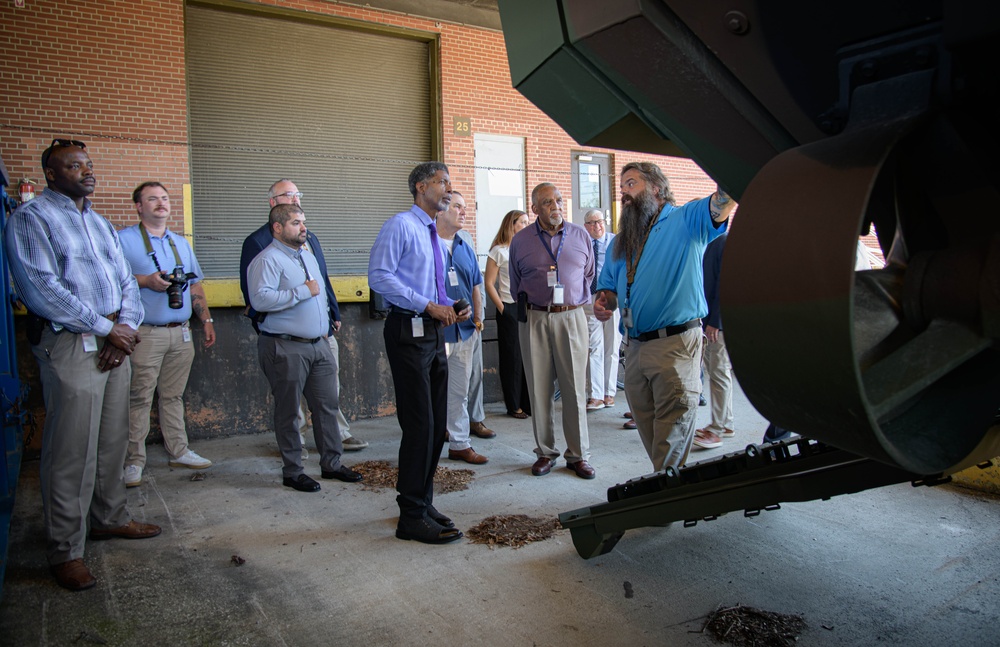 A group of approximately 12-15 professionals are receiving a guided tour or briefing at what appears to be an industrial or maintenance facility. A bearded man in a blue polo shirt is pointing toward large green industrial machinery, appearing to explain or demonstrate something to the assembled group. The setting is a covered outdoor or semi-enclosed area of a brick building with a roll-up garage door visible in the background.