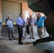 A group of approximately 12-15 professionals are receiving a guided tour or briefing at what appears to be an industrial or maintenance facility. A bearded man in a blue polo shirt is pointing toward large green industrial machinery, appearing to explain or demonstrate something to the assembled group. The setting is a covered outdoor or semi-enclosed area of a brick building with a roll-up garage door visible in the background.