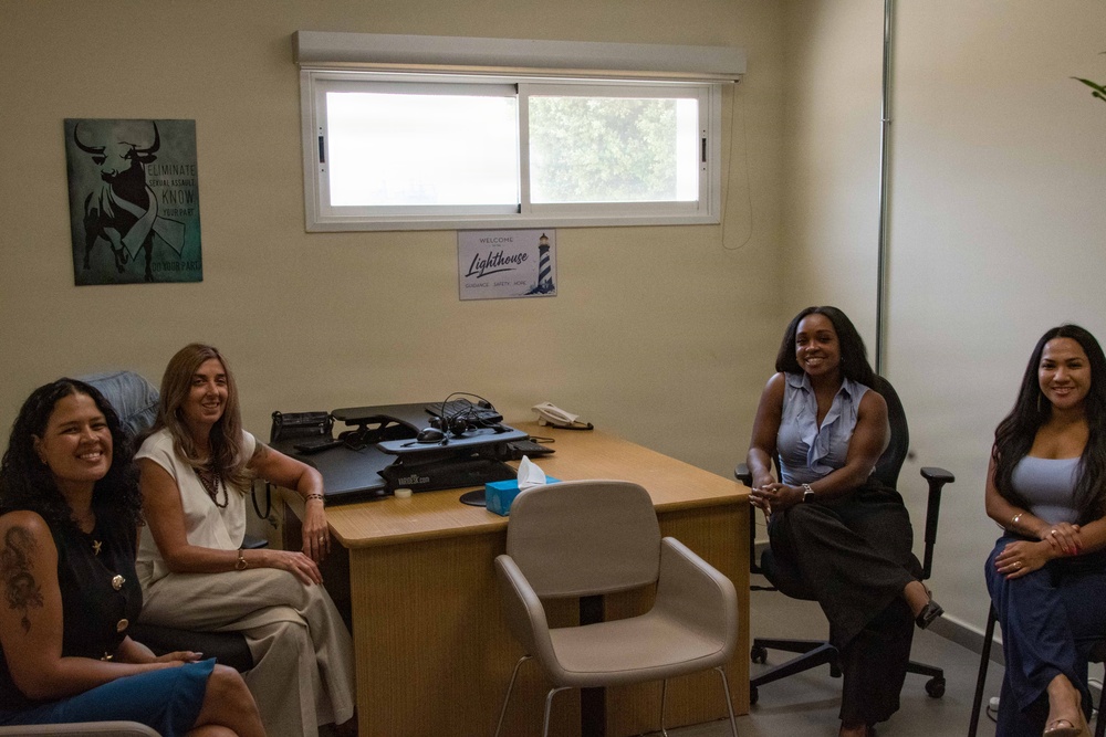 Four women are gathered in a small office setting, seated in a semi-circle arrangement around a wooden desk. The room appears to be a counseling or support office, indicated by a 'Welcome to the Lighthouse' sign on the wall near the window. All four individuals are smiling and appear to be engaged in a friendly meeting or discussion.