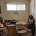 Four women are gathered in a small office setting, seated in a semi-circle arrangement around a wooden desk. The room appears to be a counseling or support office, indicated by a 'Welcome to the Lighthouse' sign on the wall near the window. All four individuals are smiling and appear to be engaged in a friendly meeting or discussion.
