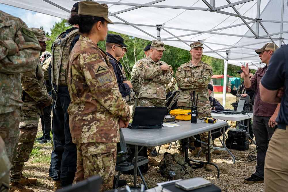 A group of military personnel from what appears to be multiple nations are gathered under a white canopy tent for a briefing or equipment demonstration. Various pieces of tactical and electronic equipment are laid out on folding tables, suggesting a field exercise or technology demonstration event. A civilian in a maroon shirt appears to be presenting or explaining something to the assembled military members.