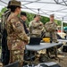 A group of military personnel from what appears to be multiple nations are gathered under a white canopy tent for a briefing or equipment demonstration. Various pieces of tactical and electronic equipment are laid out on folding tables, suggesting a field exercise or technology demonstration event. A civilian in a maroon shirt appears to be presenting or explaining something to the assembled military members.