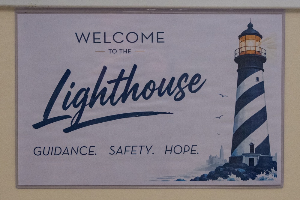 A decorative welcome sign for a place called 'The Lighthouse' mounted on a light-colored wall. The sign features a watercolor-style illustration of a black-and-white striped lighthouse with an illuminated lantern room. The tagline 'Guidance. Safety. Hope.' suggests this may be a shelter, community center, or support organization.