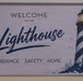 A decorative welcome sign for a place called 'The Lighthouse' mounted on a light-colored wall. The sign features a watercolor-style illustration of a black-and-white striped lighthouse with an illuminated lantern room. The tagline 'Guidance. Safety. Hope.' suggests this may be a shelter, community center, or support organization.