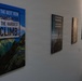 A hallway or corridor wall displays a series of motivational canvas prints arranged in a receding line. The most prominent poster features a mountain landscape (appearing to be Machu Picchu) with the quote 'The Best View Comes After the Hardest Climb.' Additional motivational posters follow along the wall, becoming less legible due to perspective and lighting.