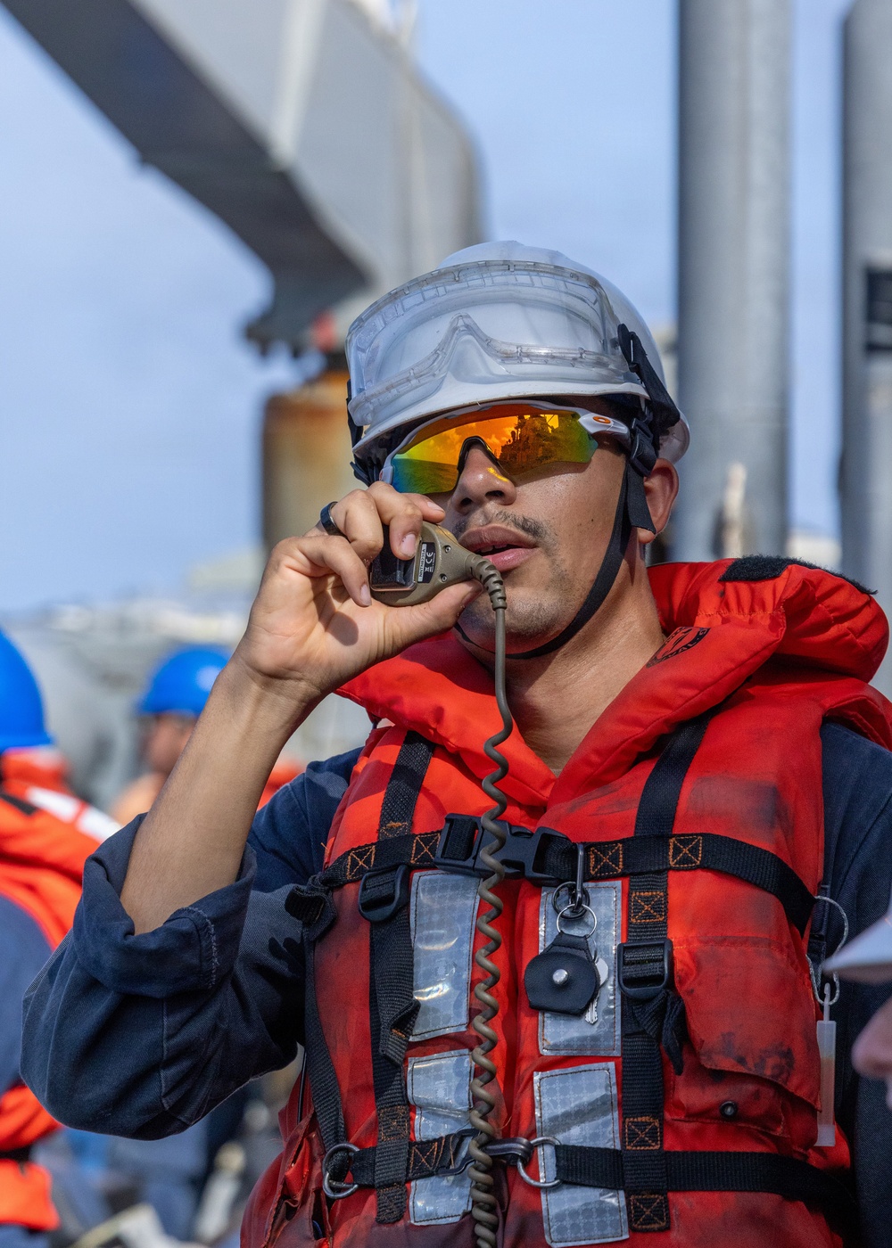 A maritime or naval worker is speaking into a corded radio handset while wearing full safety gear including a white hard hat, orange reflective life vest, and mirrored orange-tinted safety glasses. The setting appears to be the deck of a large vessel or port facility, with other personnel visible in the background also wearing safety equipment. The image captures an active communication moment during what appears to be a maritime operation or exercise.